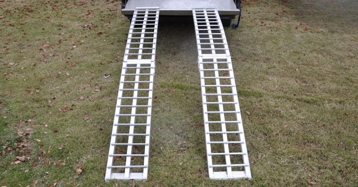 Two straight aluminium ramps extending from a trailer onto grass, ready for loading equipment.