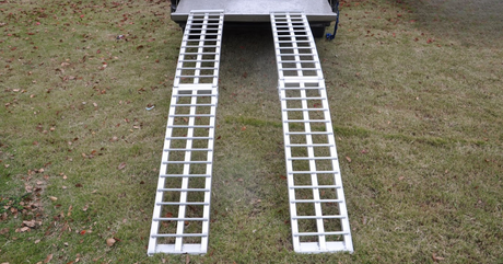 Two straight aluminium ramps extending from a trailer onto grass, ready for loading equipment.