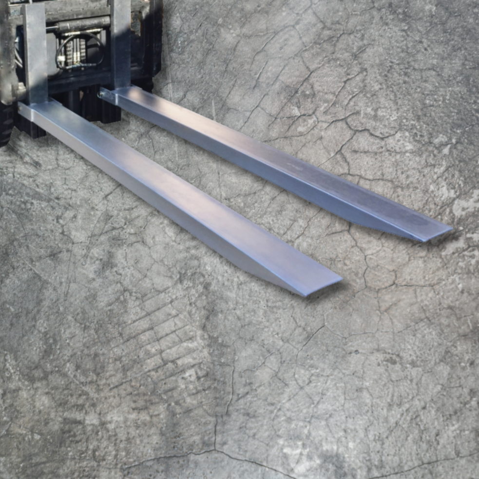 DHE Standard Duty Forklift Slippers - Ground View
A pair of steel forklift slippers resting on a cracked concrete surface, attached to a forklift, ready for use.