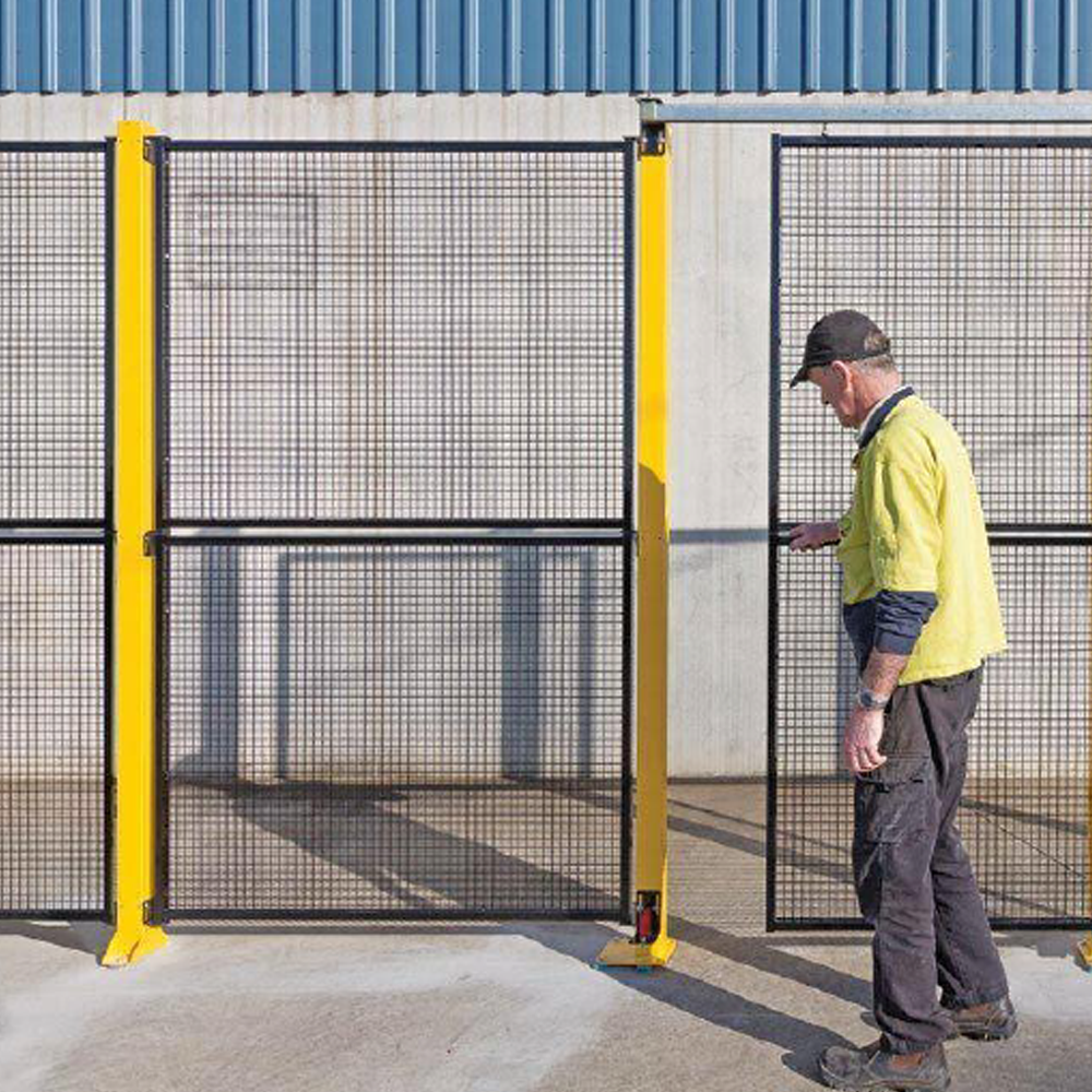 A worker wearing a high-visibility yellow jacket opens a black mesh security sliding gate with yellow steel posts, installed outside an industrial facility. The gate provides controlled entry and safety.