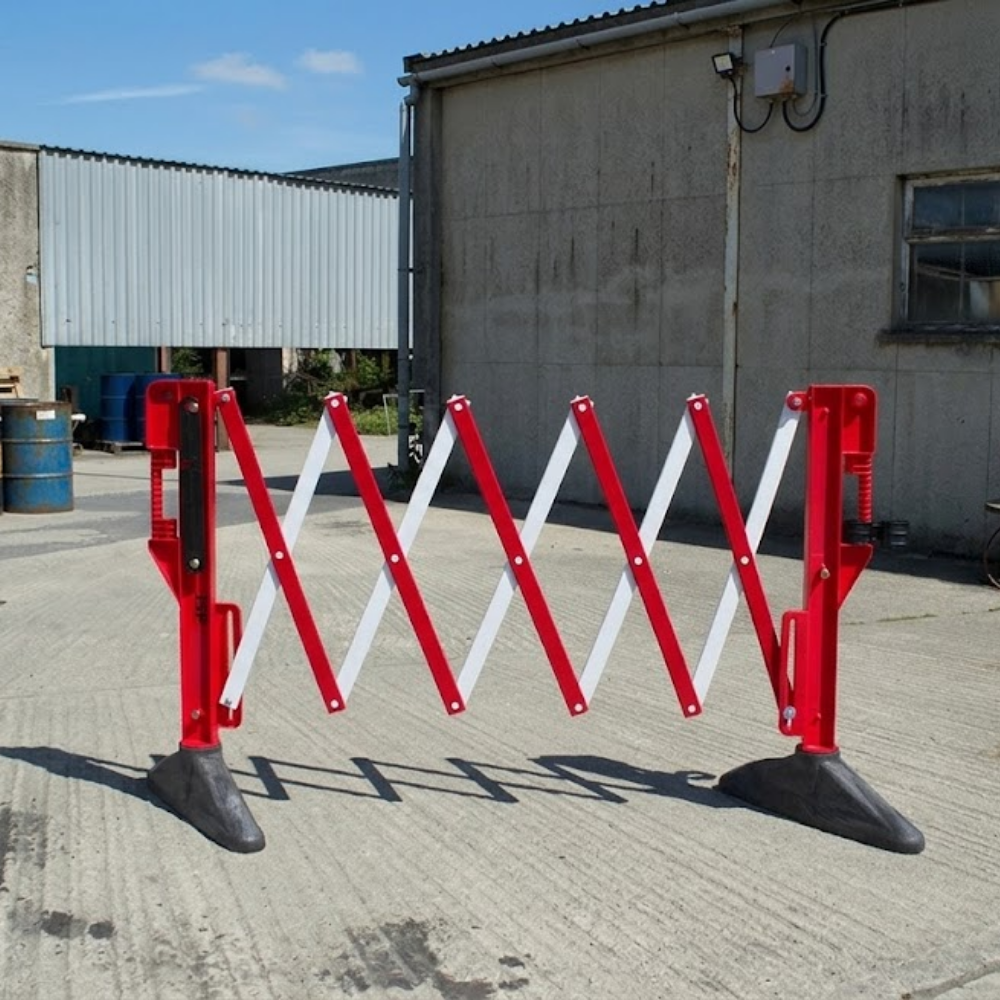 An expandable red and white plastic safety barrier with heavy black bases, set on a concrete ground outdoors.