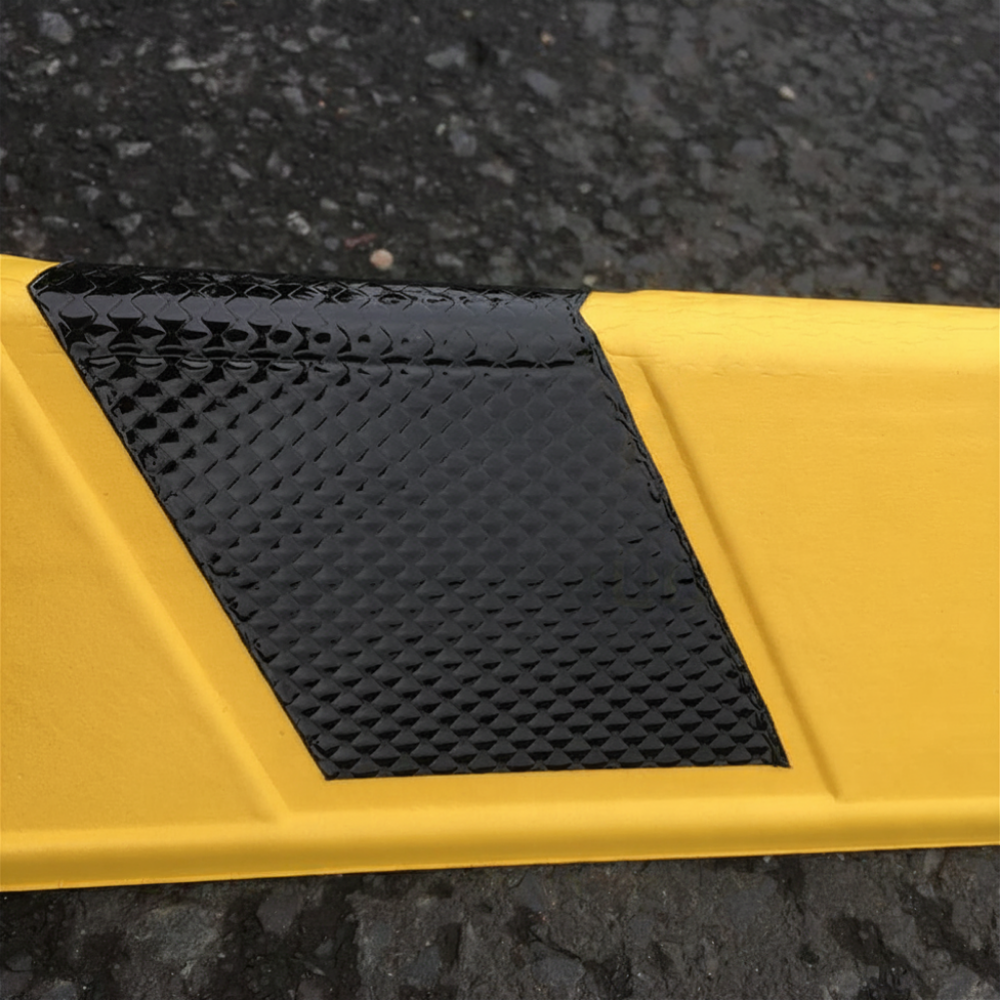 A close-up of a yellow corner guard with a textured black reflective panel on its side, photographed outdoors on dark asphalt.