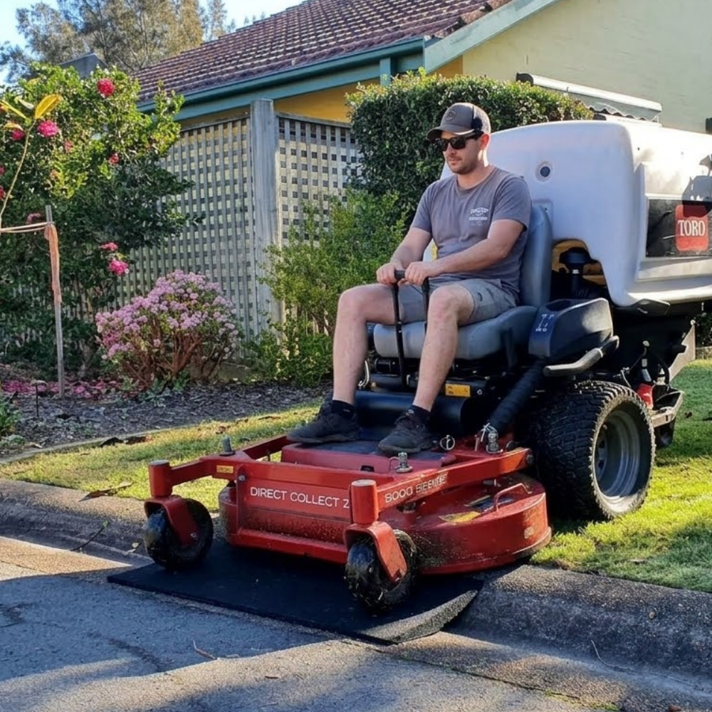 A man wearing a cap, sunglasses, and a gray t-shirt is sitting on a red and black riding lawn mower, mowing a paved driveway in a residential backyard setting.
