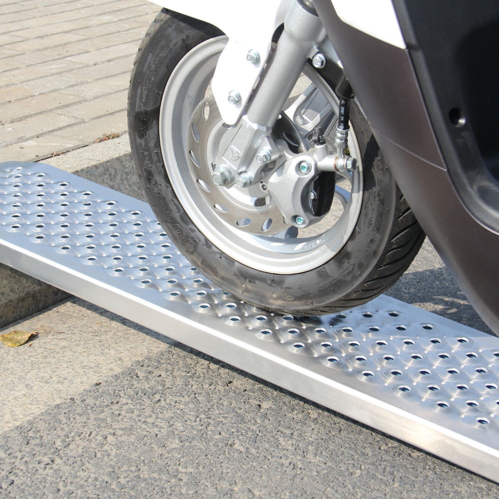 A white scooter or small motorcycle wheel is rolling onto a single aluminum ramp with a mesh surface, placed against a curb.