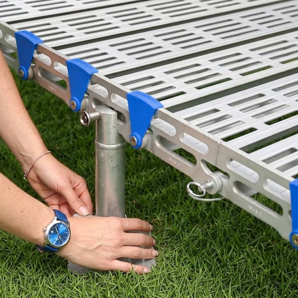 A close-up of a person's hands adjusting the metal leg of a modular outdoor floor or play mat with blue plastic clips on a green grass background.