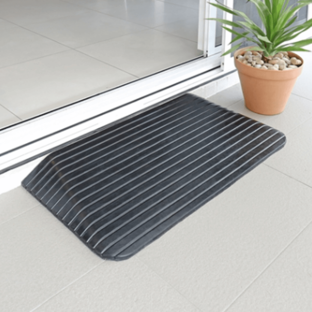 A black solid rubber wheelchair threshold door ramp with winged edges placed on a tiled floor next to a potted plant.