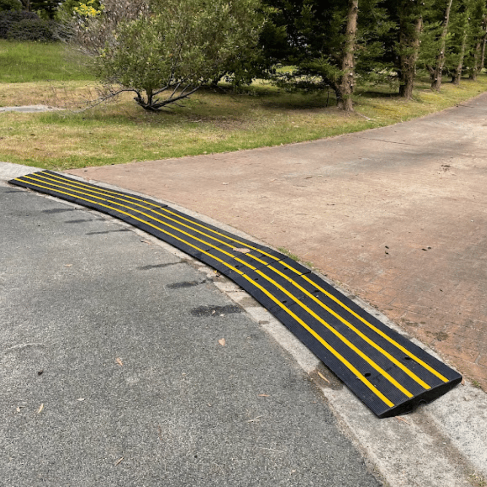 A modular black and yellow rubber driveway ramp bridges the curb of a paved driveway lined with trees.