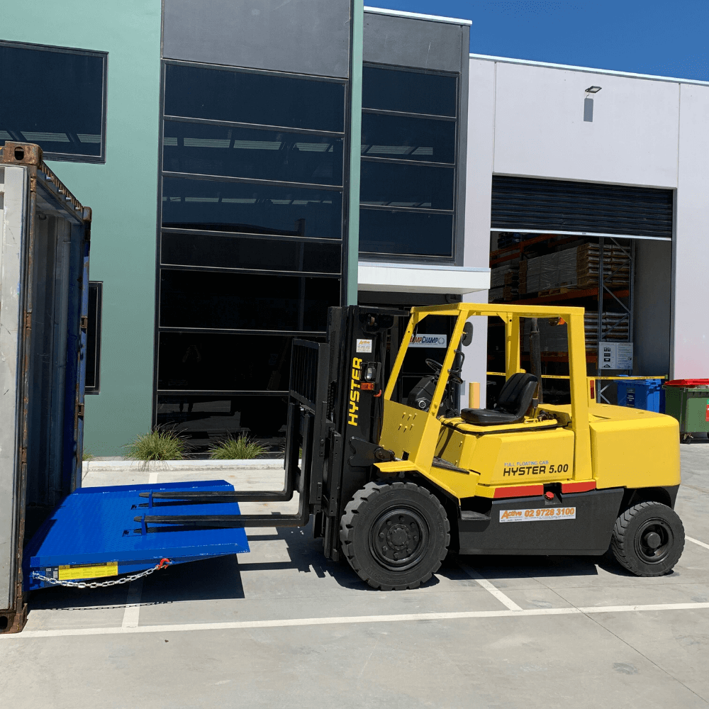 Yellow Hyster 5.00 forklift driving up a bright blue loading ramp into a shipping container, with the ramp secured by a chain and safety label visible on the side.