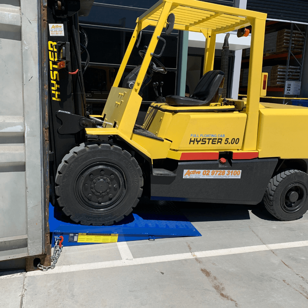 "Yellow Hyster 5.00 forklift driving up a bright blue loading ramp into a shipping container, with the ramp secured by a chain and safety label visible on the side."