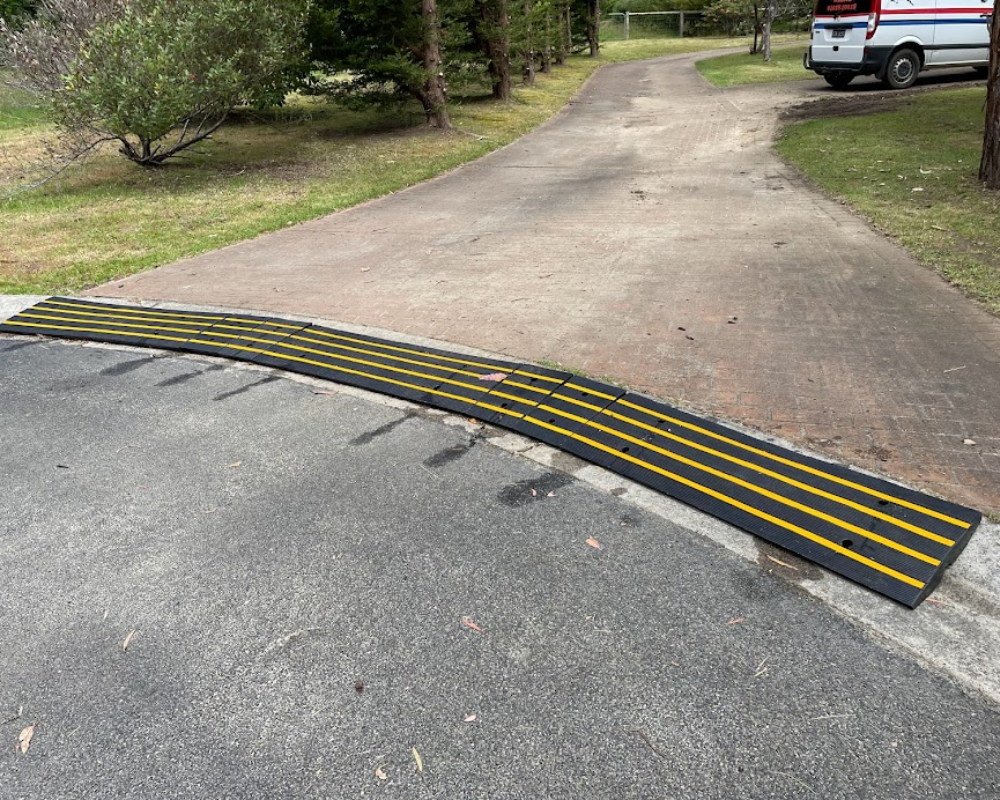 A modular black and yellow rubber driveway ramp bridges the curb of a paved driveway lined with trees and a white van.