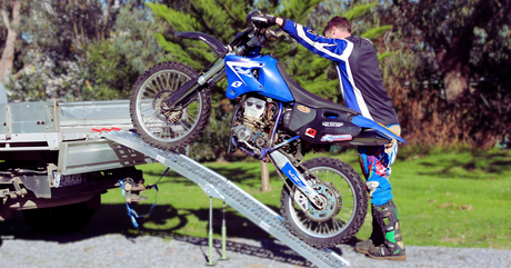 A man in motocross gear pushes a blue dirt bike up a metal loading ramp into the back of a flatbed trailer. The ramp is supported by legs for stability, and the scene is set outdoors on a sunny day with green grass and trees in the background.