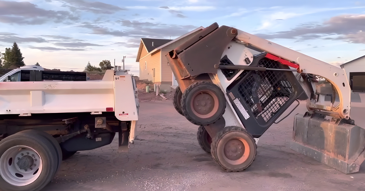 A skid steer loader lifting its front wheels off the ground while pushing against a dump truck in a construction setting, with a suburban house and a sunset sky in the background.