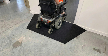 A person in a power wheelchair uses a black rubber threshold ramp to enter a doorway.