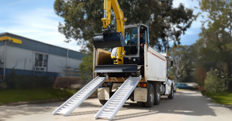 Excavator driving onto a truck using a pair of heavy-duty aluminium machinery ramps.