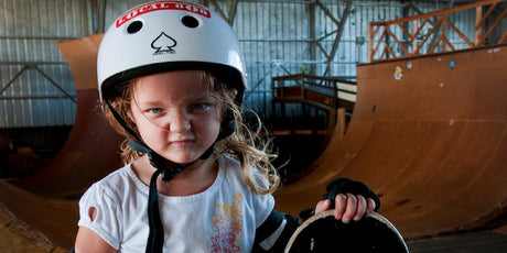 A young girl wearing helmet and holding a skateboard at a skateboard park