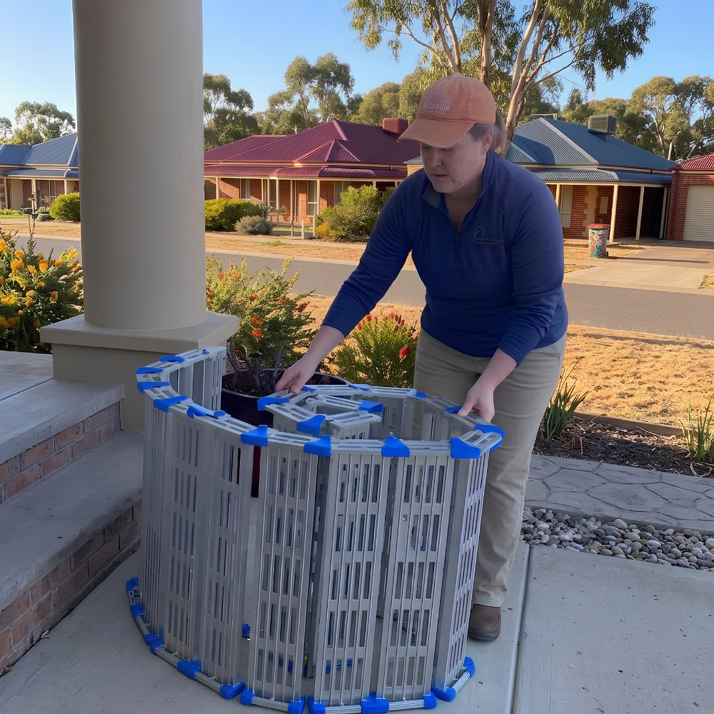 A woman in a blue long-sleeve shirt and khaki pants is assembling a circular silver aluminum pet playpen with blue plastic connectors on a concrete porch in front of a house.