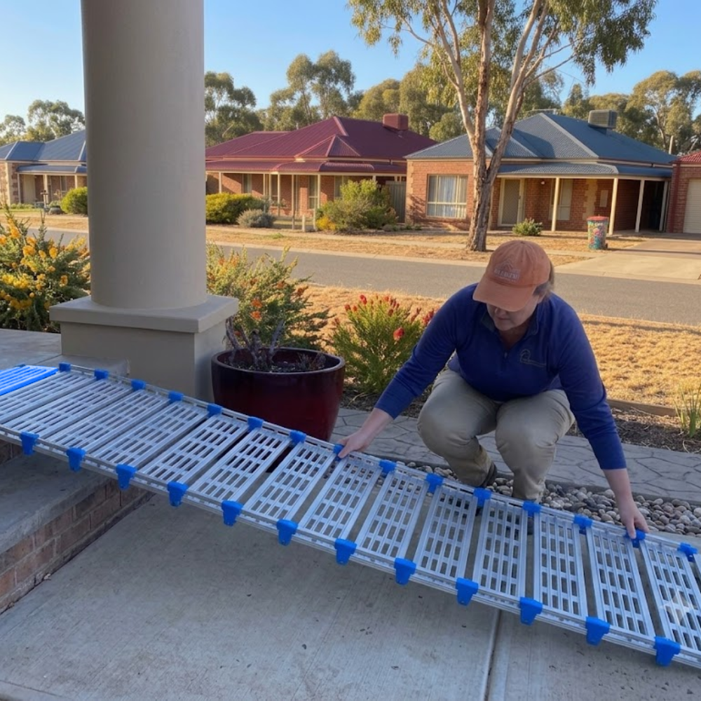 A person crouches on a concrete porch while holding a long, silver metal ramp with blue plastic connectors, with a suburban street and houses in the background.