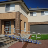A metal wheelchair ramp with blue grip pads and silver metal handrails, positioned on the driveway in front of a residential house.