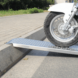 Aluminum ramp with mesh surface bridging a curb, with a motorcycle/scooter wheel starting to roll onto it.