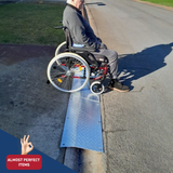 Elderly man in a wheelchair using a portable aluminium kerb ramp on a street, labelled as an “Almost Perfect Item.”