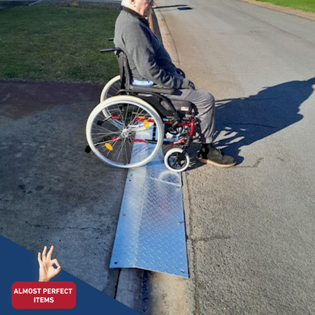 Elderly man in a wheelchair using a portable aluminium kerb ramp on a street, labelled as an “Almost Perfect Item.”