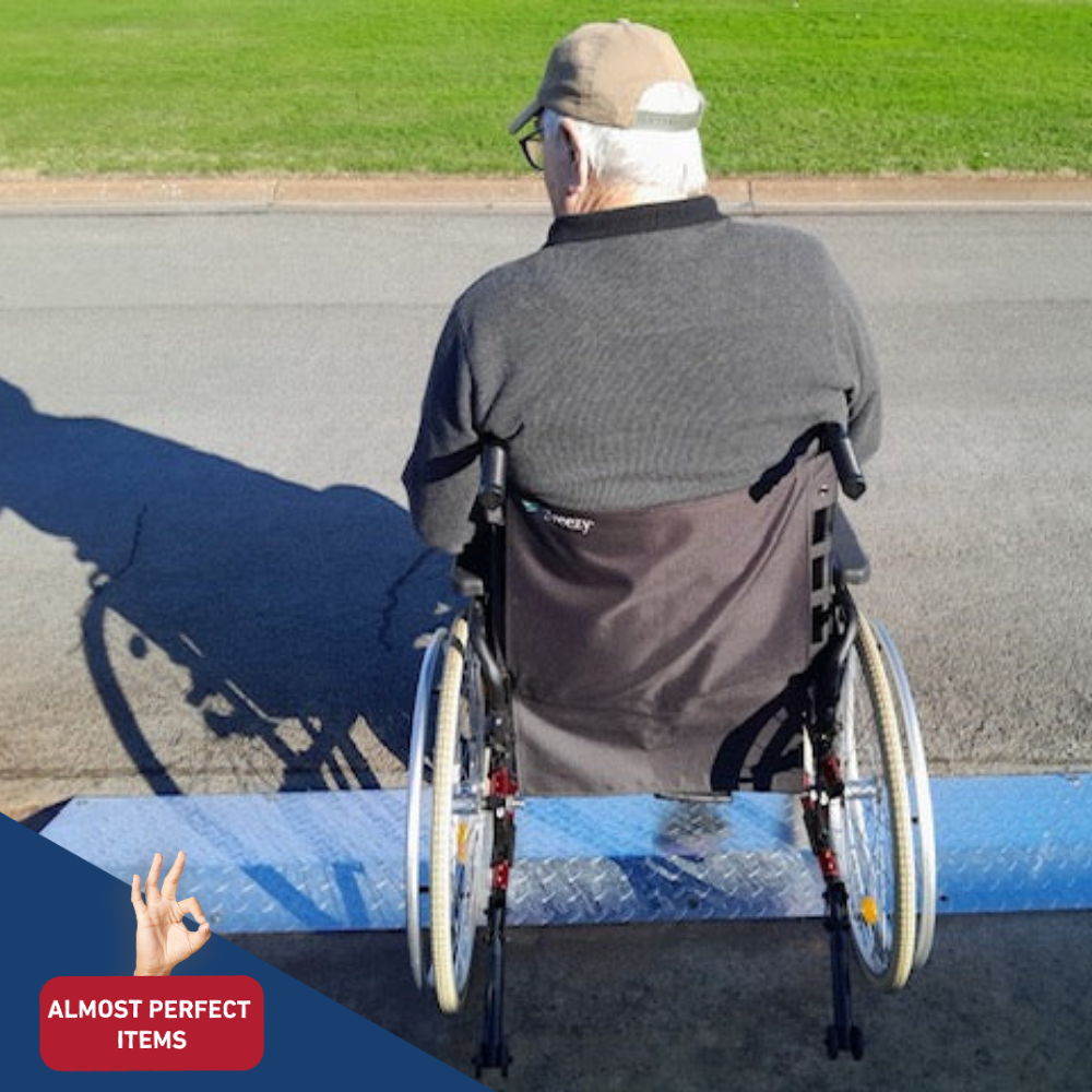 Back of an elderly man in a wheelchair using a portable aluminium kerb ramp on a street, labelled as an “Almost Perfect Item.”