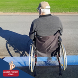 Back of an elderly man in a wheelchair using a portable aluminium kerb ramp on a street, labelled as an “Almost Perfect Item.”