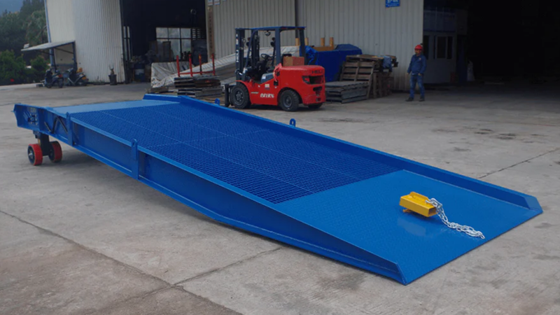 Blue loading ramp with a yellow chain on a concrete floor, with a forklift and person in the background.