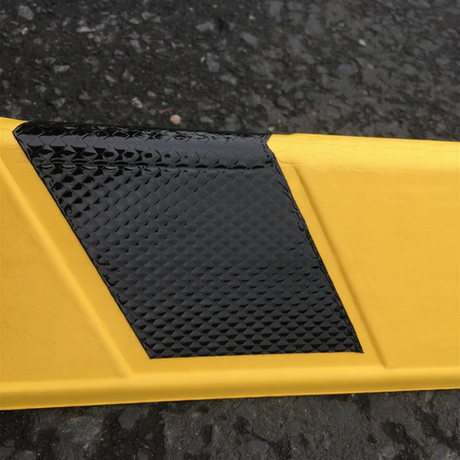 A close-up of a yellow corner guard with a textured black reflective panel on its side, photographed outdoors on dark asphalt.