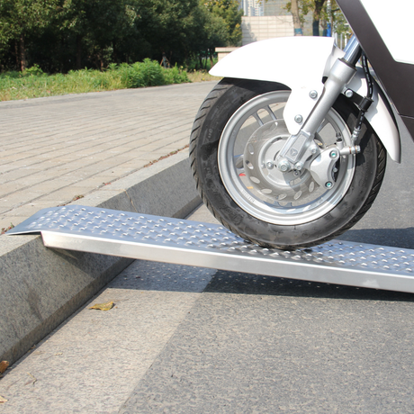 A white scooter or small motorcycle wheel is rolling onto a single aluminum ramp with a mesh surface, placed against a curb.