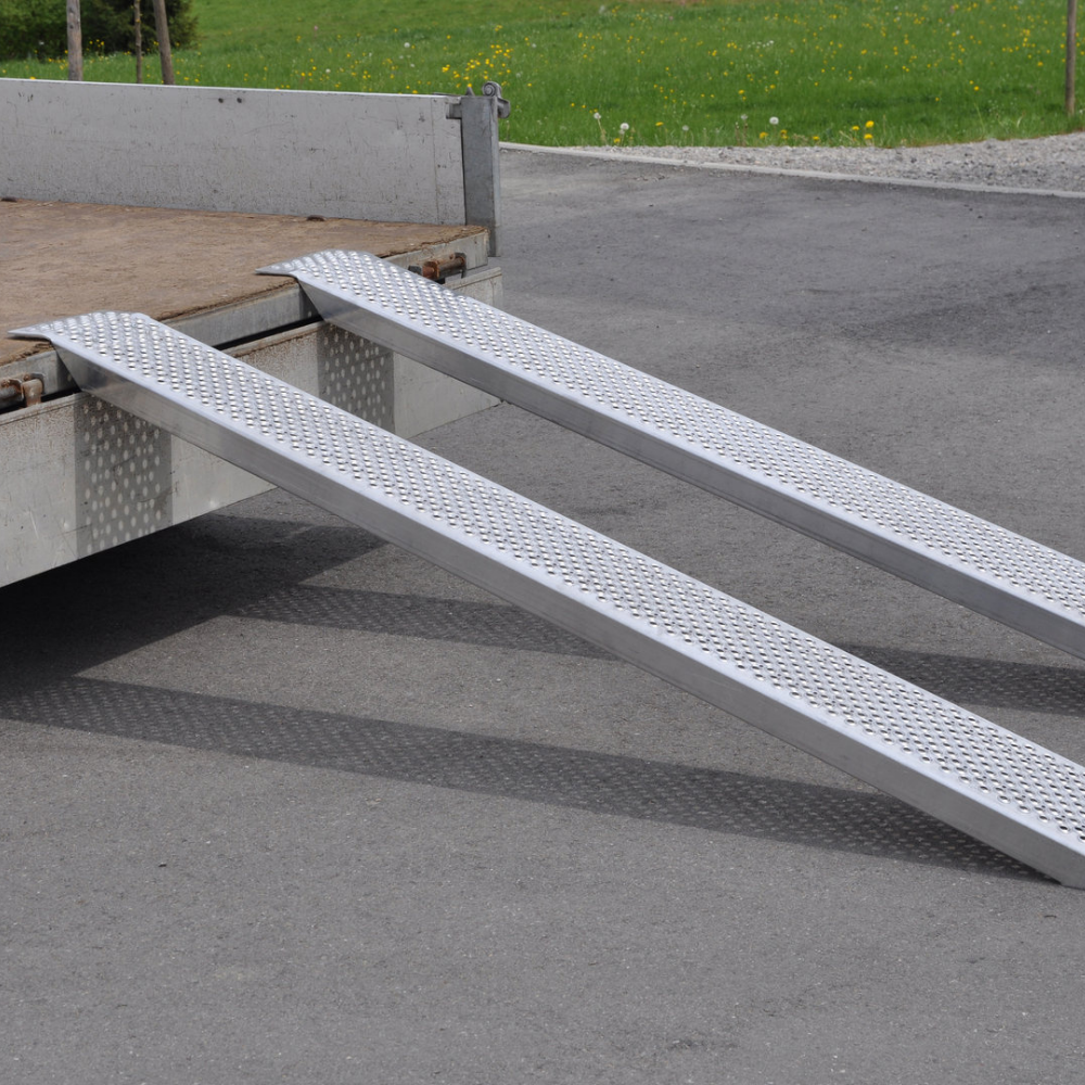 A pair of straight aluminium ramps with a perforated mesh surface are set up, resting on the back edge of a flatbed trailer.
