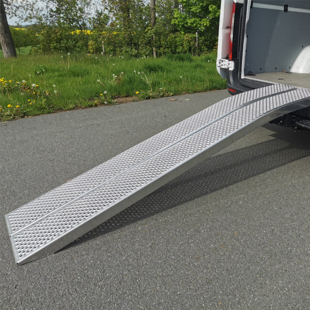 A wide, single aluminum loading ramp with a mesh, non-slip surface extending from the open back of a white van to the asphalt road.