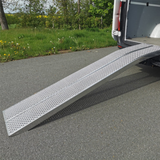 A wide, single aluminum loading ramp with a mesh, non-slip surface extending from the open back of a white van to the asphalt road.