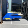 Forklift driving up a bright blue loading ramp into a shipping container, with the ramp secured by a chain and safety label visible on the side.
