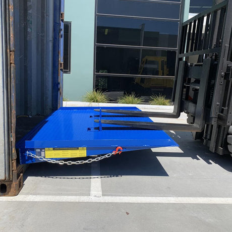 Forklift driving up a bright blue loading ramp into a shipping container, with the ramp secured by a chain and safety label visible on the side.
