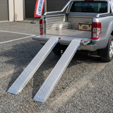 Two truck ramps extended from a pickup truck bed with a visible brand logo in the background.