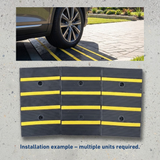 A car tyre rolls onto a black rubber kerb ramp with yellow safety stripes. Multiple interlocking units are shown below.
