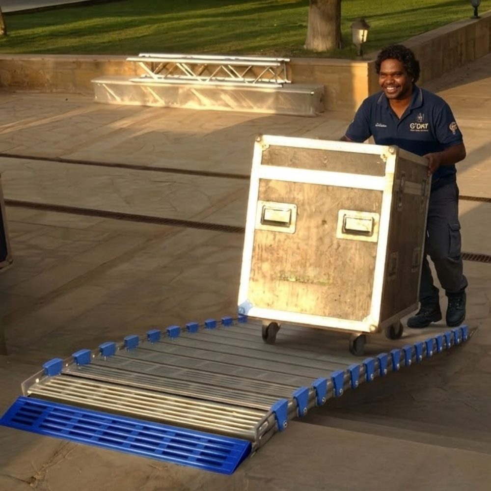 A man in a navy blue uniform pushes a large wooden equipment case up a metal ramp with blue plastic side supports outdoors on a paved walkway.