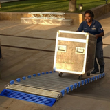 A man in a navy blue uniform pushes a large wooden equipment case up a metal ramp with blue plastic side supports outdoors on a paved walkway.