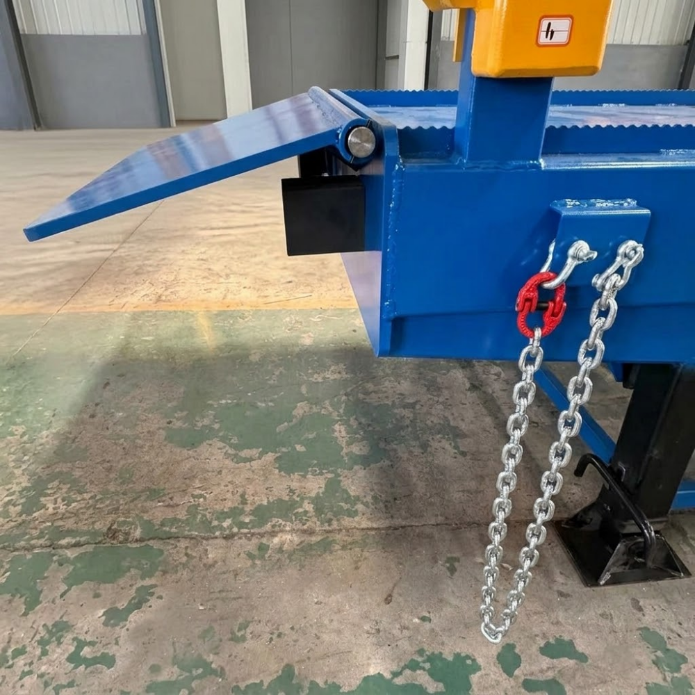 A close-up of a blue forklift yard ramp, likely a compactor or press, with a yellow control box, a blue inclined chute, and a silver chain hanging from a red hook. The machine is photographed in a warehouse or factory setting on a concrete floor.