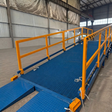 A blue metal inclined yard ramp with bright yellow safety railings, shown inside a large warehouse or industrial building.