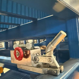 Closeup of a hydraulic control unit with a red handwheel and metal lever mounted beneath a blue grated forklift loading ramp.
