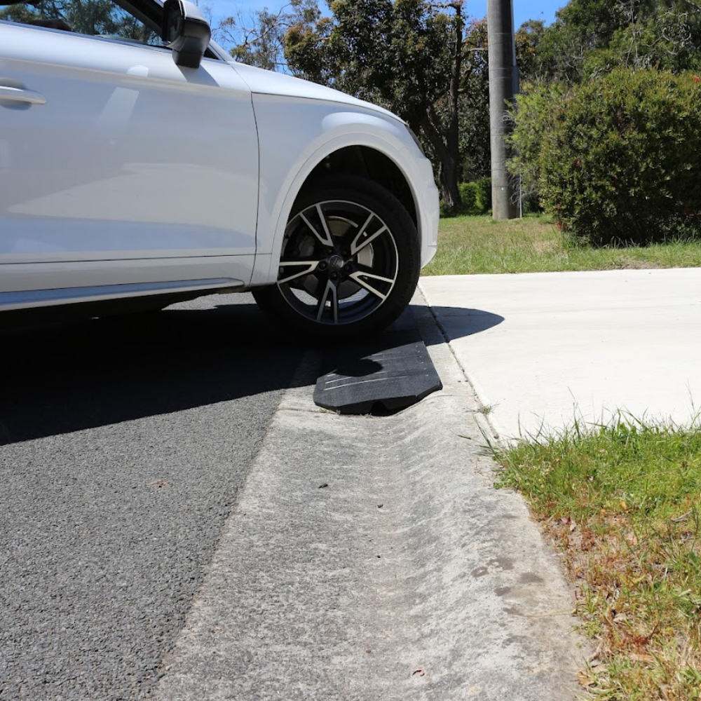 A side-profile view of a white SUV driving over the curb using the ramp. This image illustrates the smooth transition the ramp provides for a vehicle entering a residential driveway.