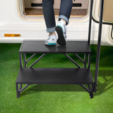 Person using a black step stool to climb into a vehicle on a grassy area.